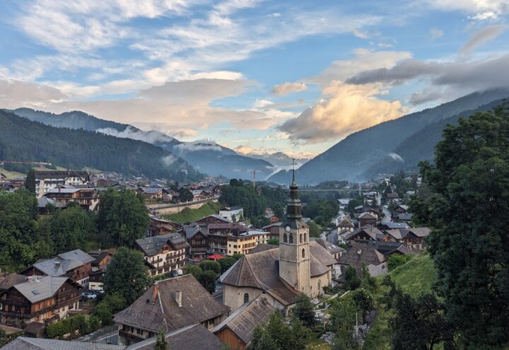 Uitzicht over Morzine met een kerk, houten huizen en beboste bergen onder een wolkenlucht