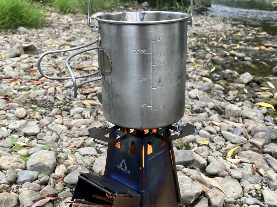 Review: FireTower By Arnaud 1 Metalen kookpot boven een brandende FireTower op een rotsachtige rivieroever
