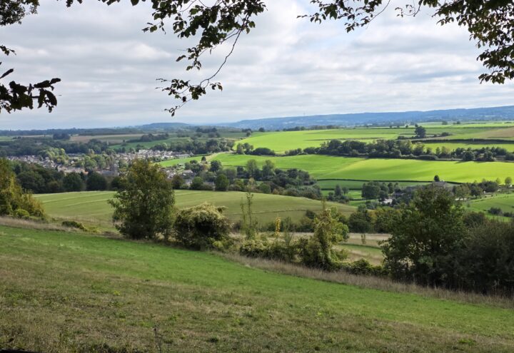 Uitzicht over groene velden, een dorp en beboste heuvels onder een bewolkte lucht