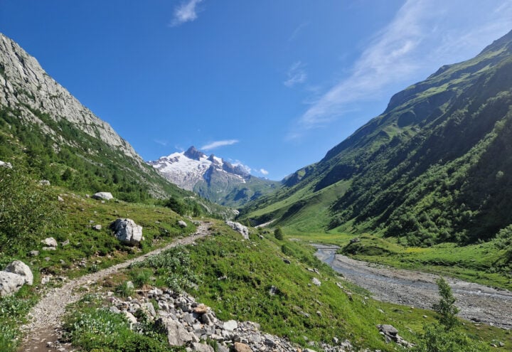 Bergdal met een rotsachtig wandelpad, groene hellingen en besneeuwde bergtoppen onder een blauwe lucht