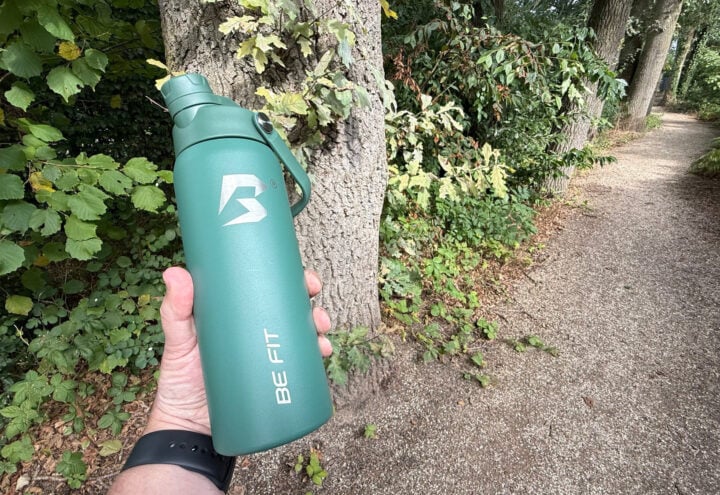 Groene BE FIT Swigger in een hand naast een onverhard wandelpad in het bos