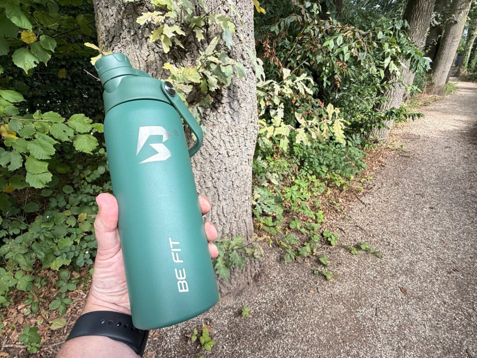 BE FIT Swigger review 1 Groene BE FIT Swigger in een hand naast een onverhard wandelpad in het bos