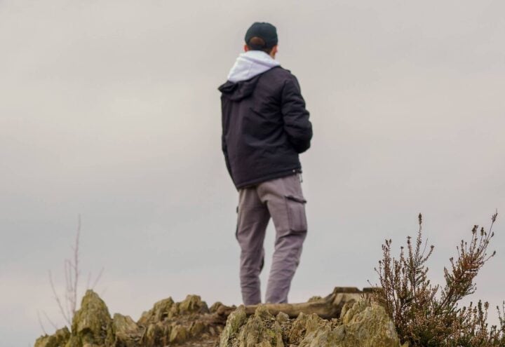 Wandelaar in donkere jas staat op een rotsachtige heuvel onder een bewolkte lucht