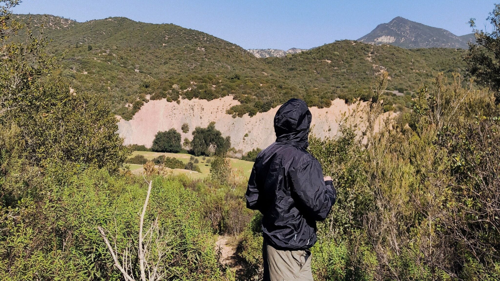 Wat is een softshell jas, en wanneer kies je een hardshell? 2 Wandelaar in een donkere jas op een bergpad bij droog, helder weer