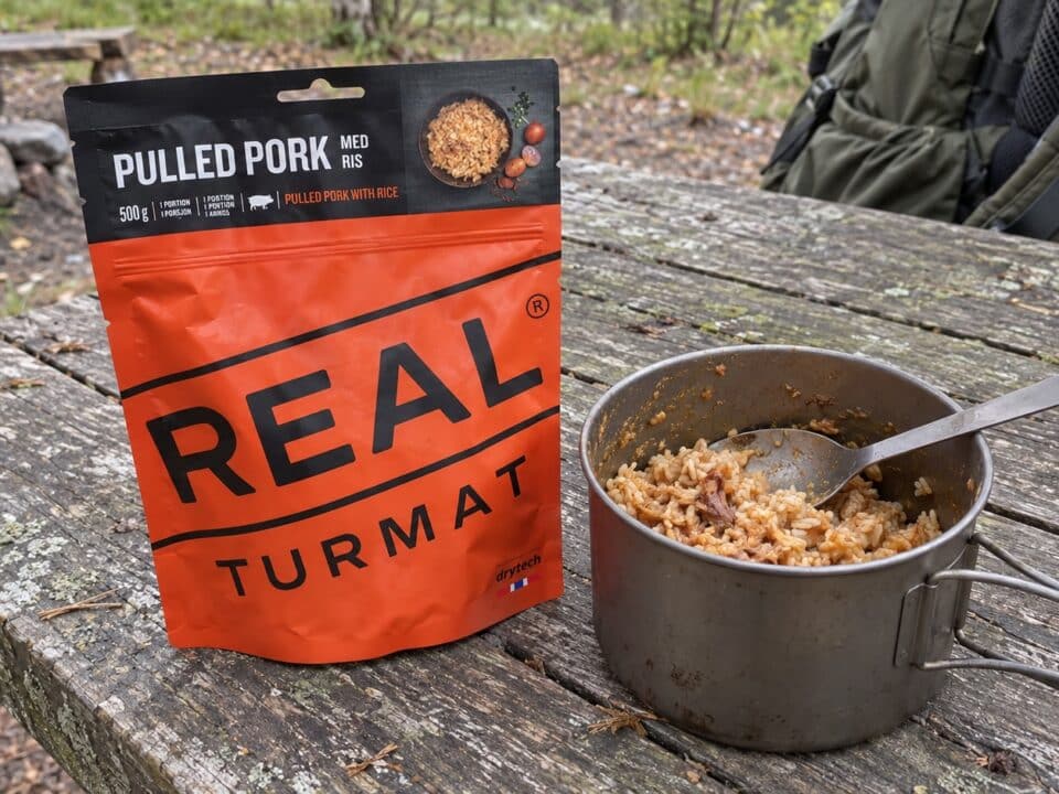 Zak Real Turmat Pulled Pork met rijst naast een metalen kookpot op een houten kampeertafel