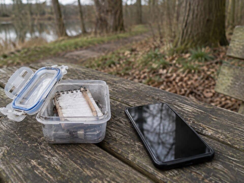 Open kunststof doos met logboek en smartphone op een houten picknicktafel bij een vijver