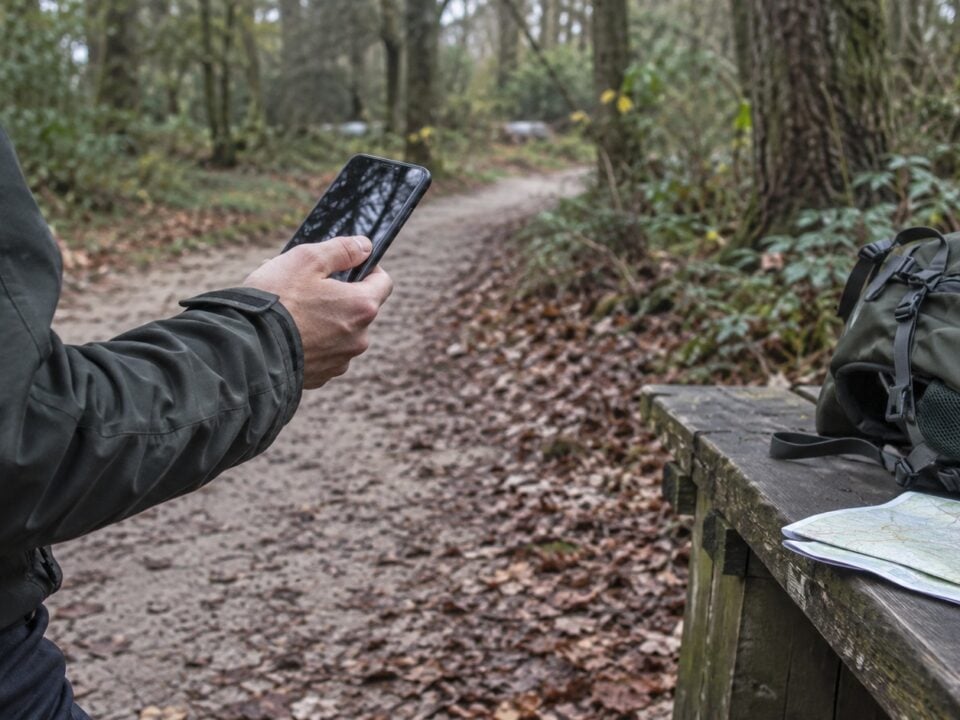 Wandelaar gebruikt een smartphone naast een rugzak, kaart en powerbank op een houten bankje in het bos