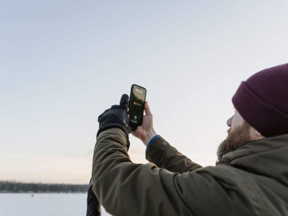 Telefoon in de kou: zo houd je je batterij bruikbaar 1 Man met muts gebruikt een smartphone bij een besneeuwd landschap